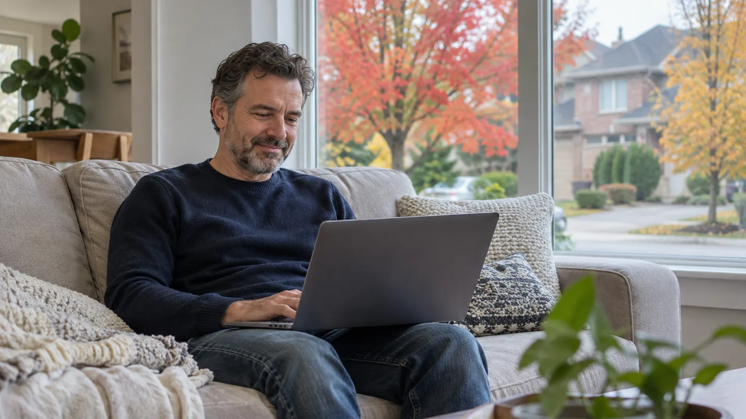A man in his forties using a laptop on a sofa at home in Canada, autumn maples outside the window